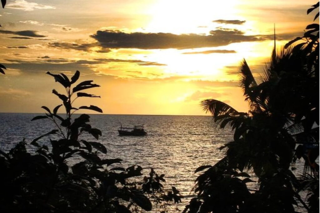 Pôr do sol dourado sobre o mar em Freedom beach Phuket, com silhueta de vegetação tropical e um barco ao fundo