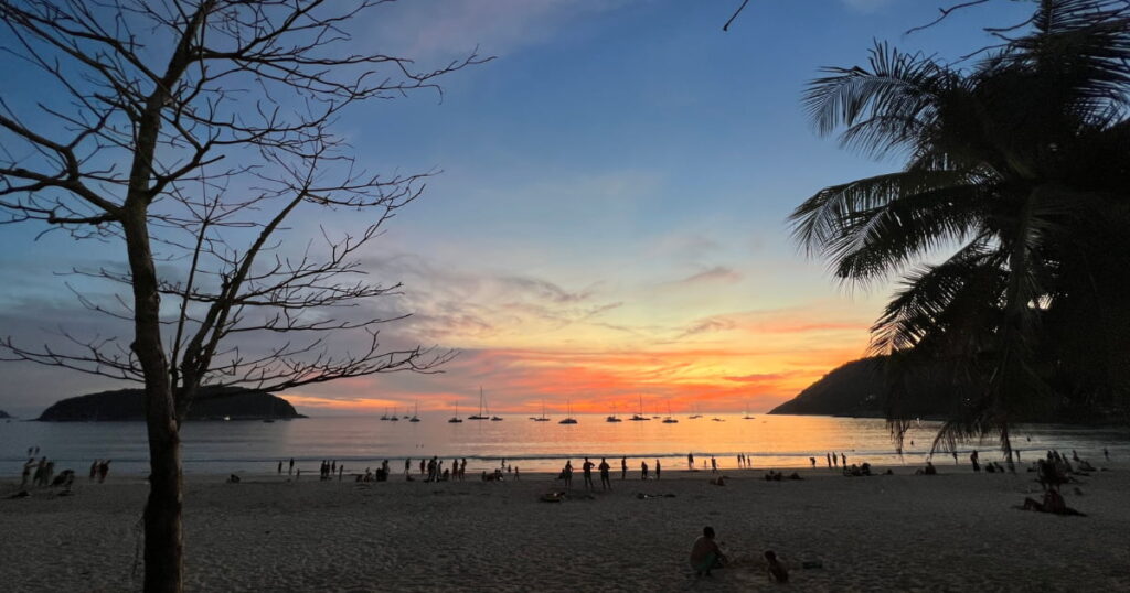 Pôr do sol laranja e rosa sobre a praia de Phuket, com turistas na areia, veleiros ancorados no mar e palmeiras ao redor