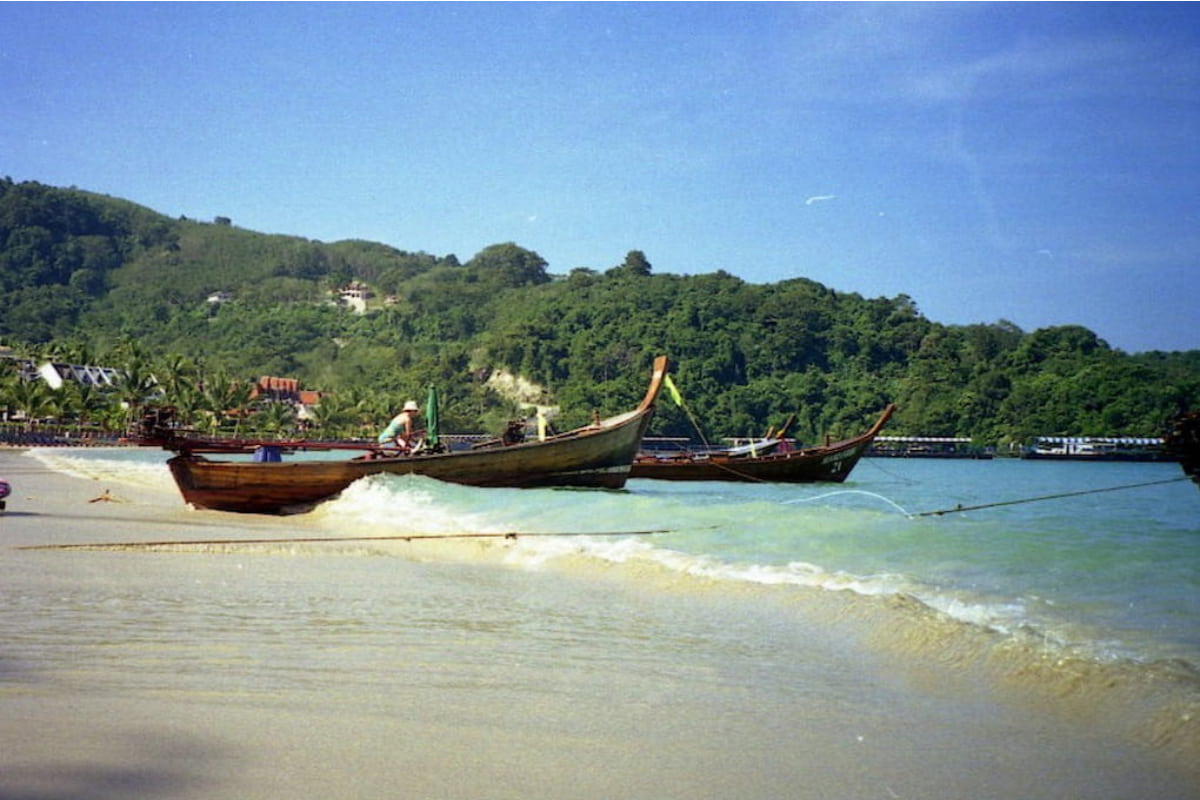 Barcos de madeira ancorados na areia da Patong Beach, com vegetação verde nas colinas ao fundo, em Phuket, Tailândia