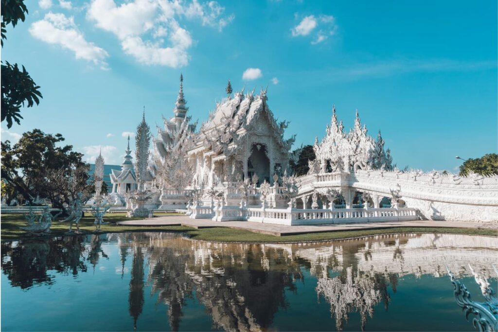 Templo Branco de Chiang Rai refletido no lago, com estrutura toda branca e detalhes elaborados sob céu azul, na Tailândia